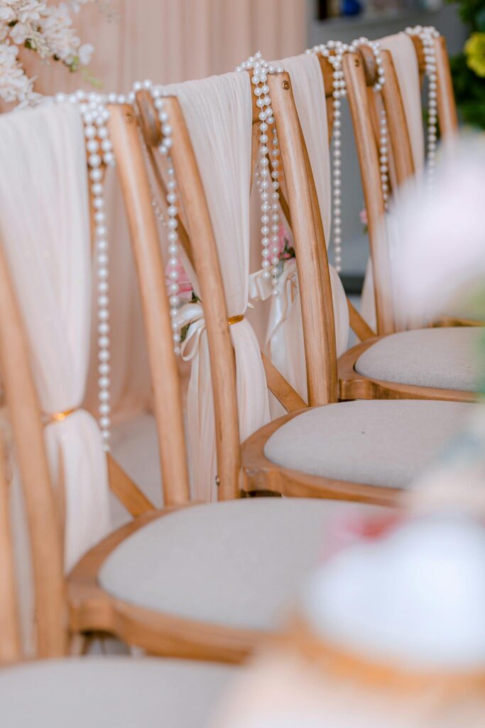 Rows of elegant wooden chairs adorned with pearl strings and floral arrangements at a wedding ceremony.