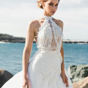 A stunning bride in an elegant white gown stands gracefully by the ocean.