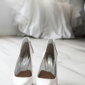 Stylish white bridal shoes positioned in front of a flowing wedding dress indoors.