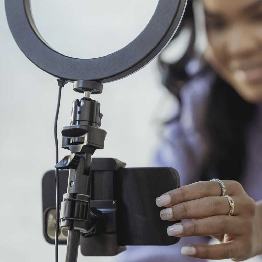 Cheerful young African American female blogger in stylish sweater smiling while setting up camera of smartphone attached to tripod with ring light before recording vlog