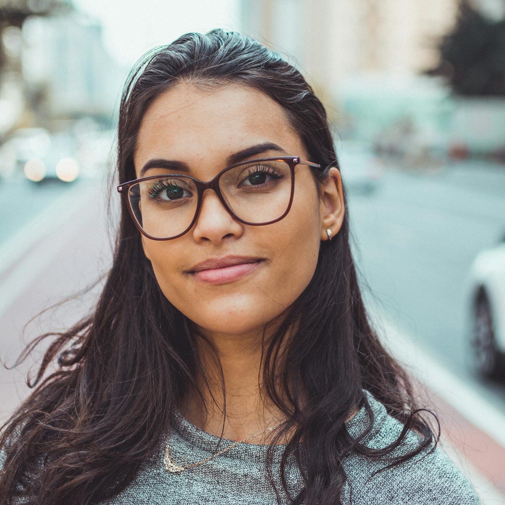 A confident woman wearing glasses smiles subtly while standing on an urban street with blurred background.
