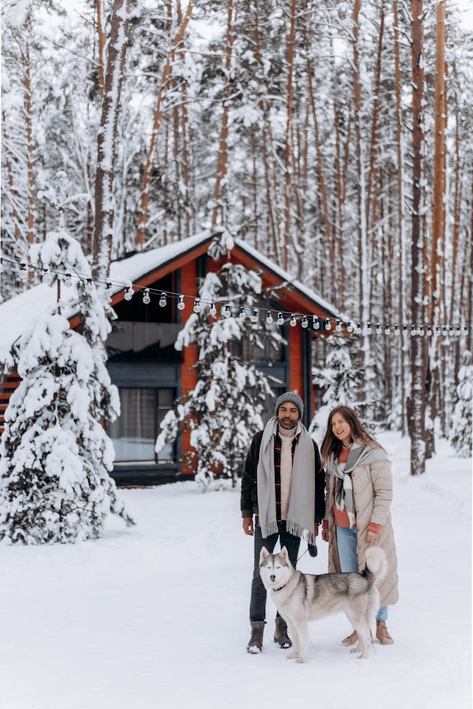 A joyful couple with their husky enjoys a winter holiday in a snowy forest by a cozy cabin.