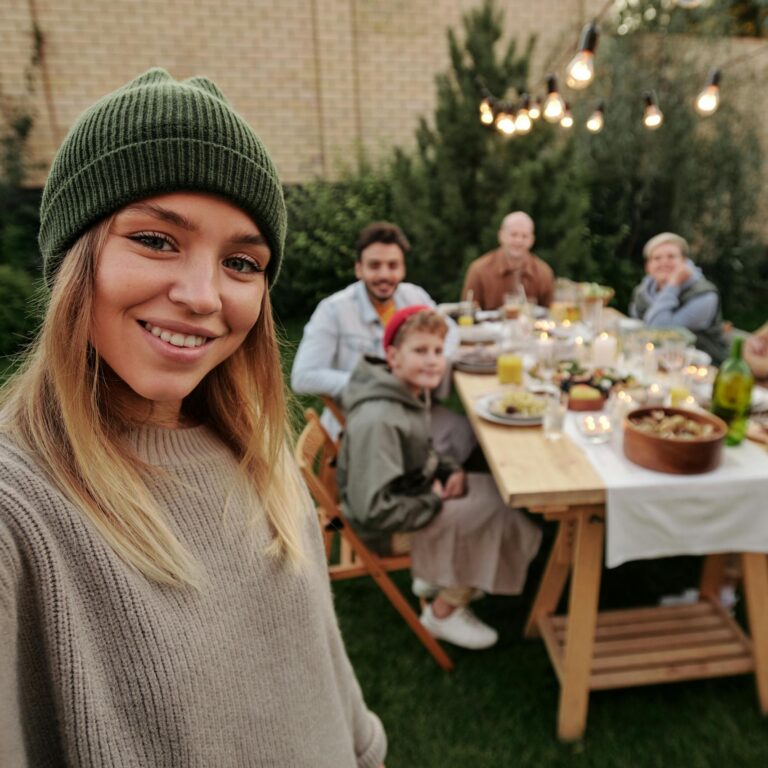 A joyful family dinner outdoors, captured with a smiling woman in the foreground taking a selfie.