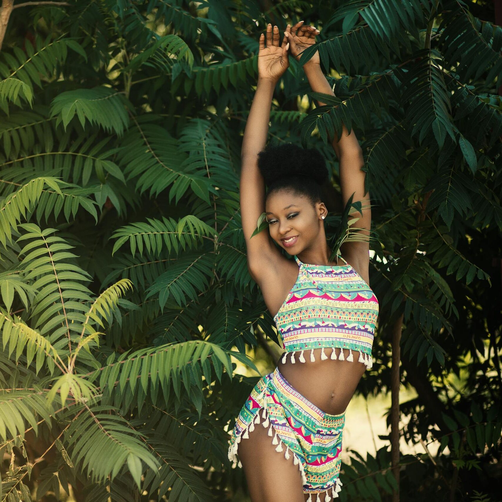 A joyful young woman posing in colorful attire amidst lush greenery outdoors.