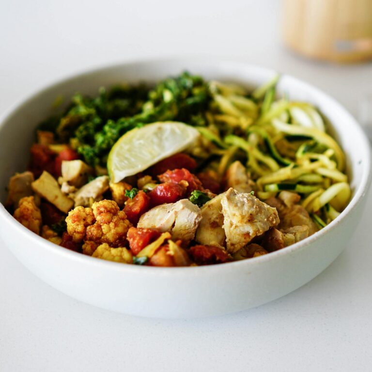 A nutritious bowl containing zoodles, roasted cauliflower, chicken, and a lemon slice, perfect for healthy eating.