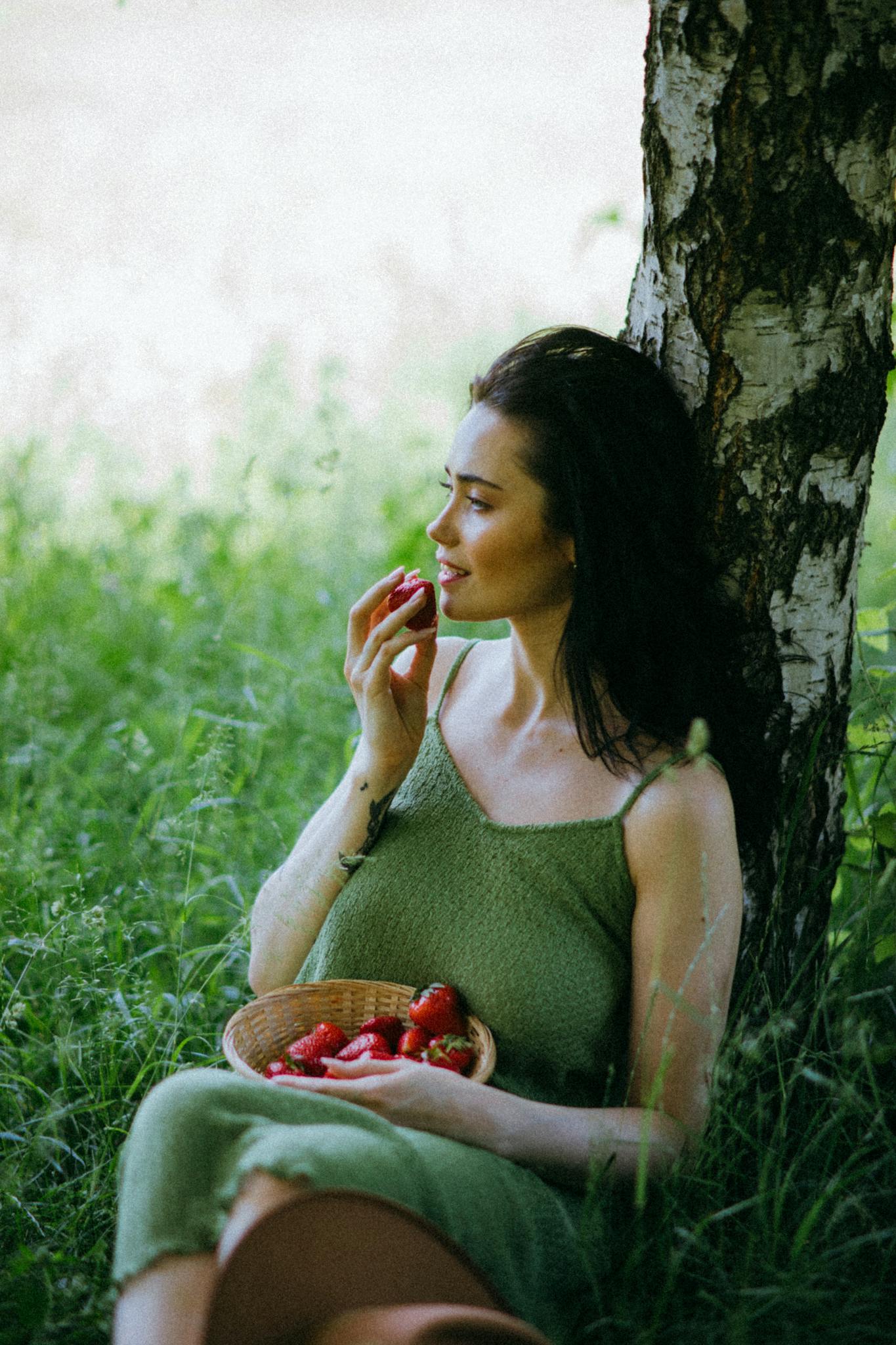 A woman in a green dress enjoys strawberries by a birch tree on a sunny day.