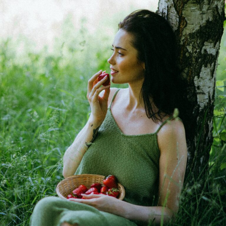 A woman in a green dress enjoys strawberries by a birch tree on a sunny day.