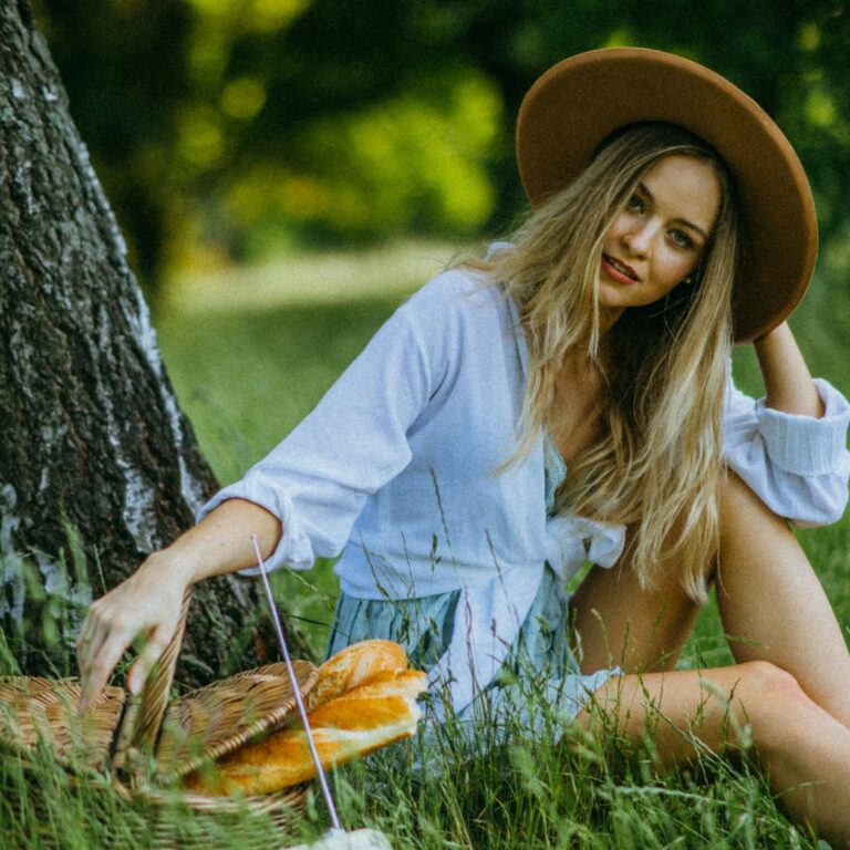 A woman in a hat enjoys a summer picnic, sitting by a tree in a lush green park.