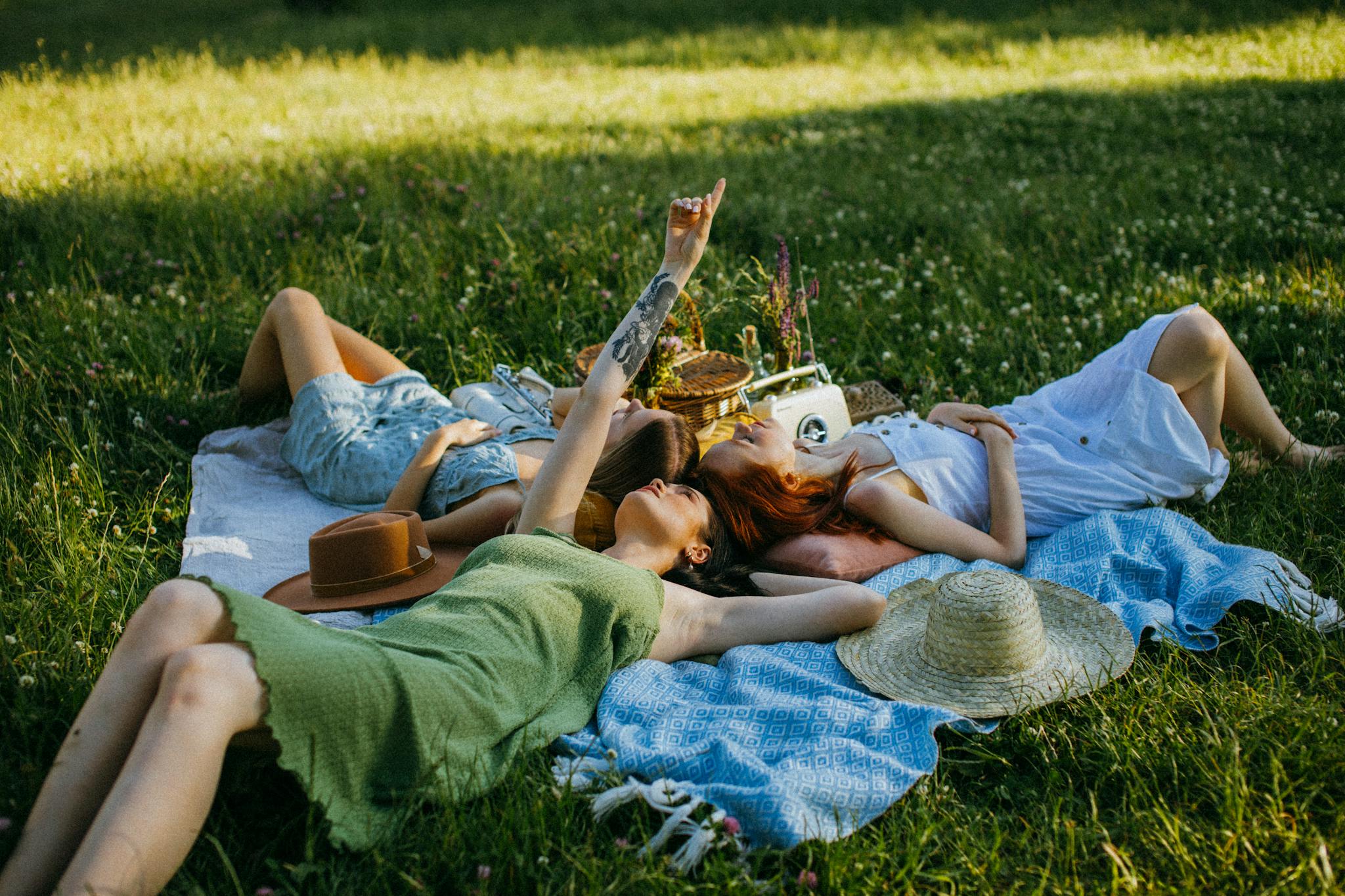 Group of women enjoying a relaxing picnic in a sunny park, lying on a blanket, surrounded by greenery.