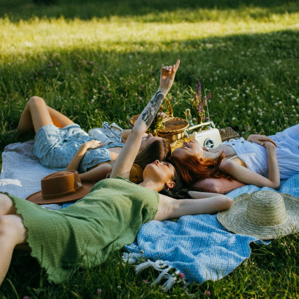 Group of women enjoying a relaxing picnic in a sunny park, lying on a blanket, surrounded by greenery.