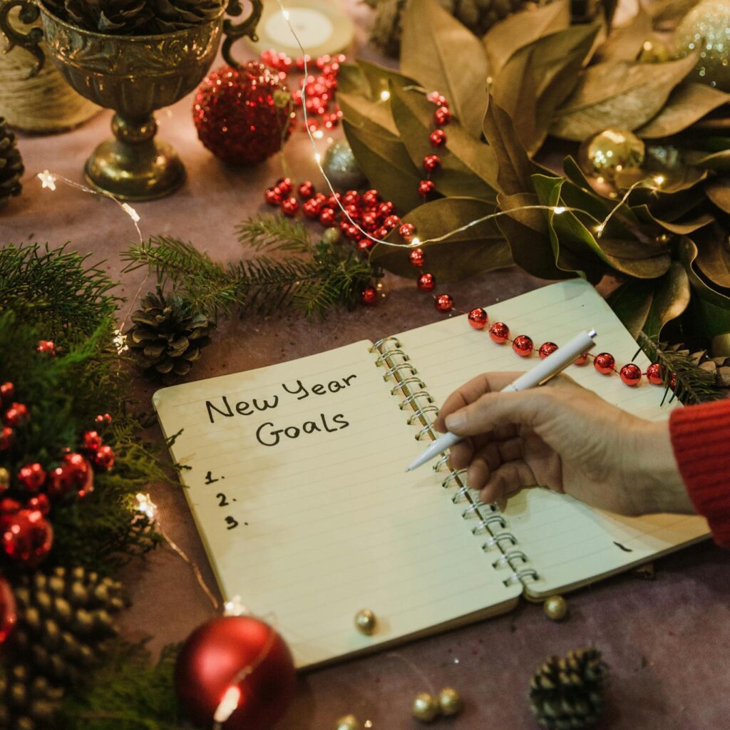 Hand writing New Year goals in a festive notebook surrounded by Christmas decorations and fairy lights indoors.