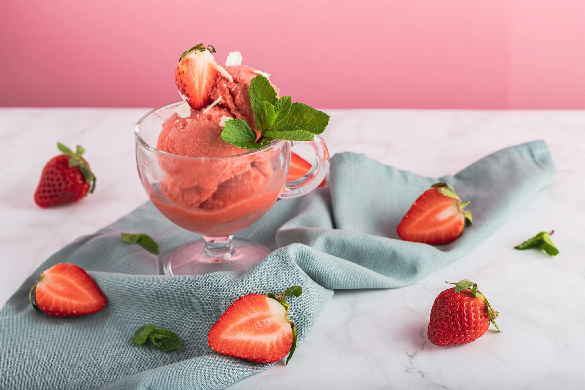 Refreshing strawberry sorbet in a glass, garnished with mint and fresh sliced strawberries on a pink background.