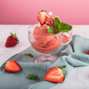 Refreshing strawberry sorbet in a glass, garnished with mint and fresh sliced strawberries on a pink background.