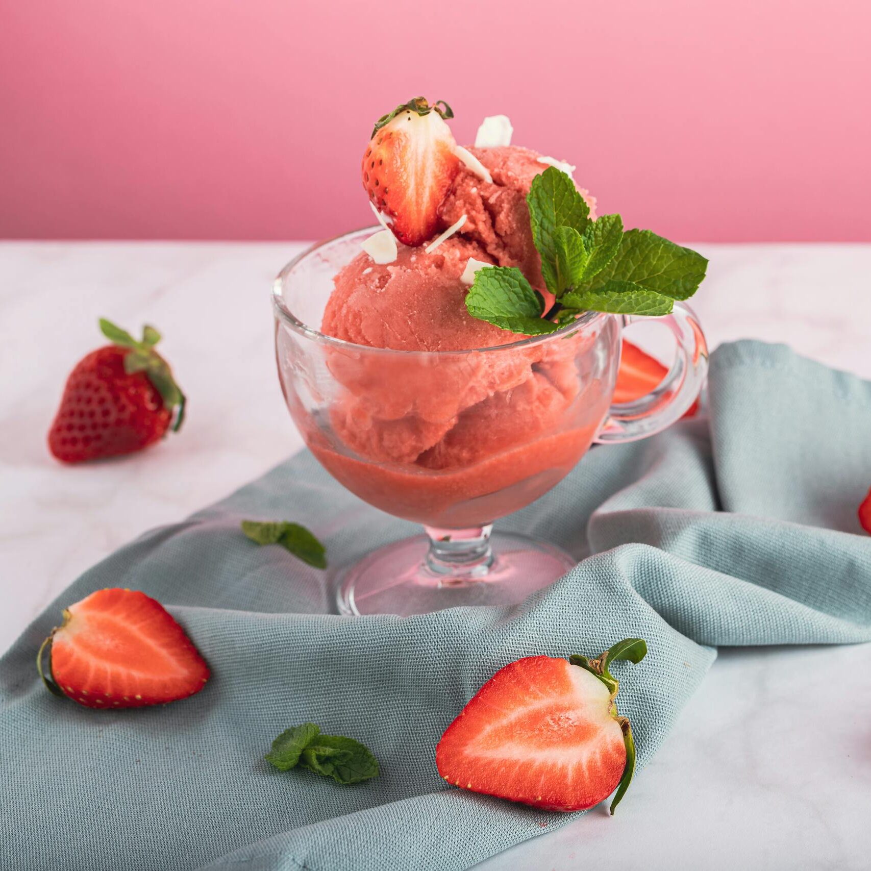 Refreshing strawberry sorbet in a glass, garnished with mint and fresh sliced strawberries on a pink background.
