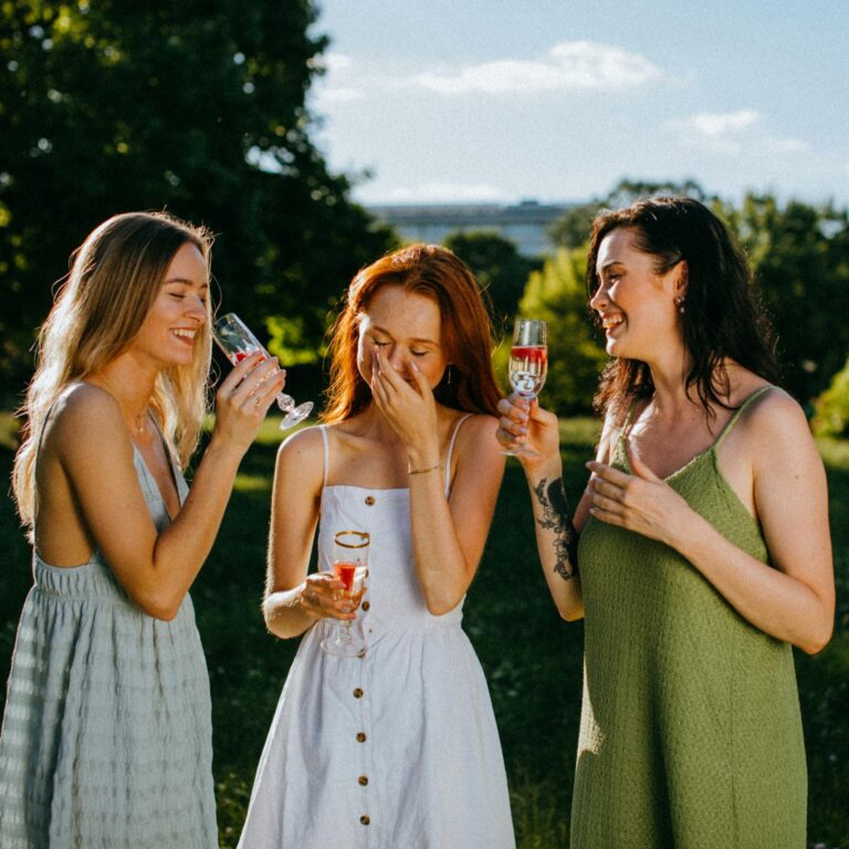 Three women enjoying a sunny day with drinks, celebrating friendship outdoors.
