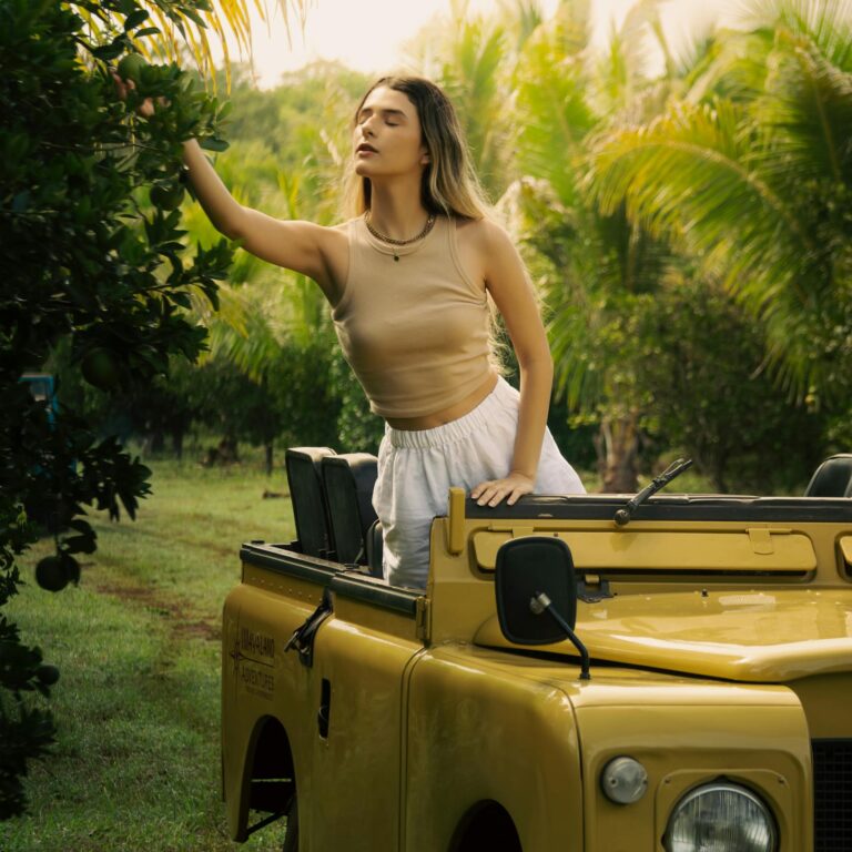 Woman beside a vintage yellow jeep reaching out towards leaves in a tropical setting.
