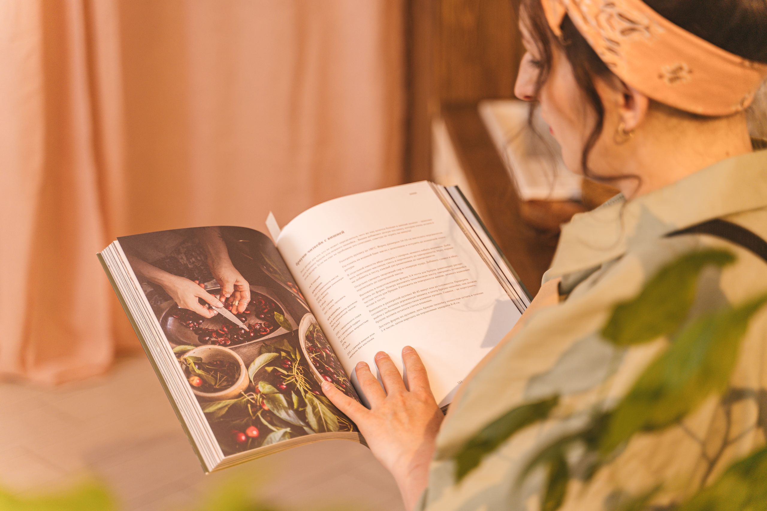 A woman reading a cookbook with colorful images and recipes in a warm indoor setting.