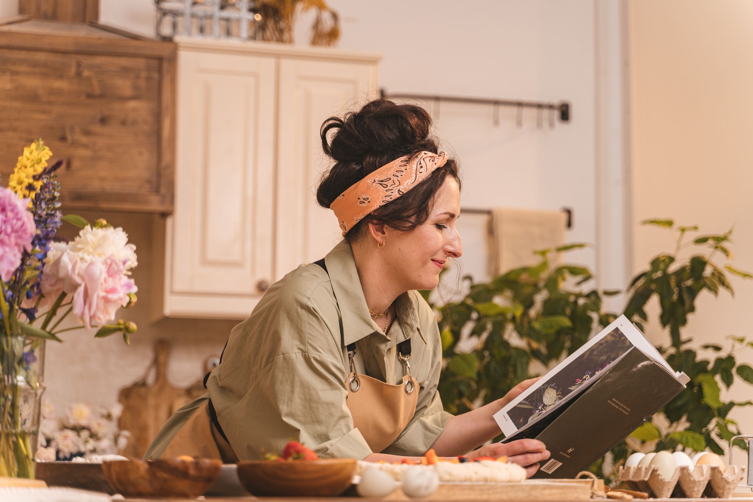 Woman in kitchen reading a cookbook, surrounded by flowers and cooking ingredients. Warm and homely atmosphere.