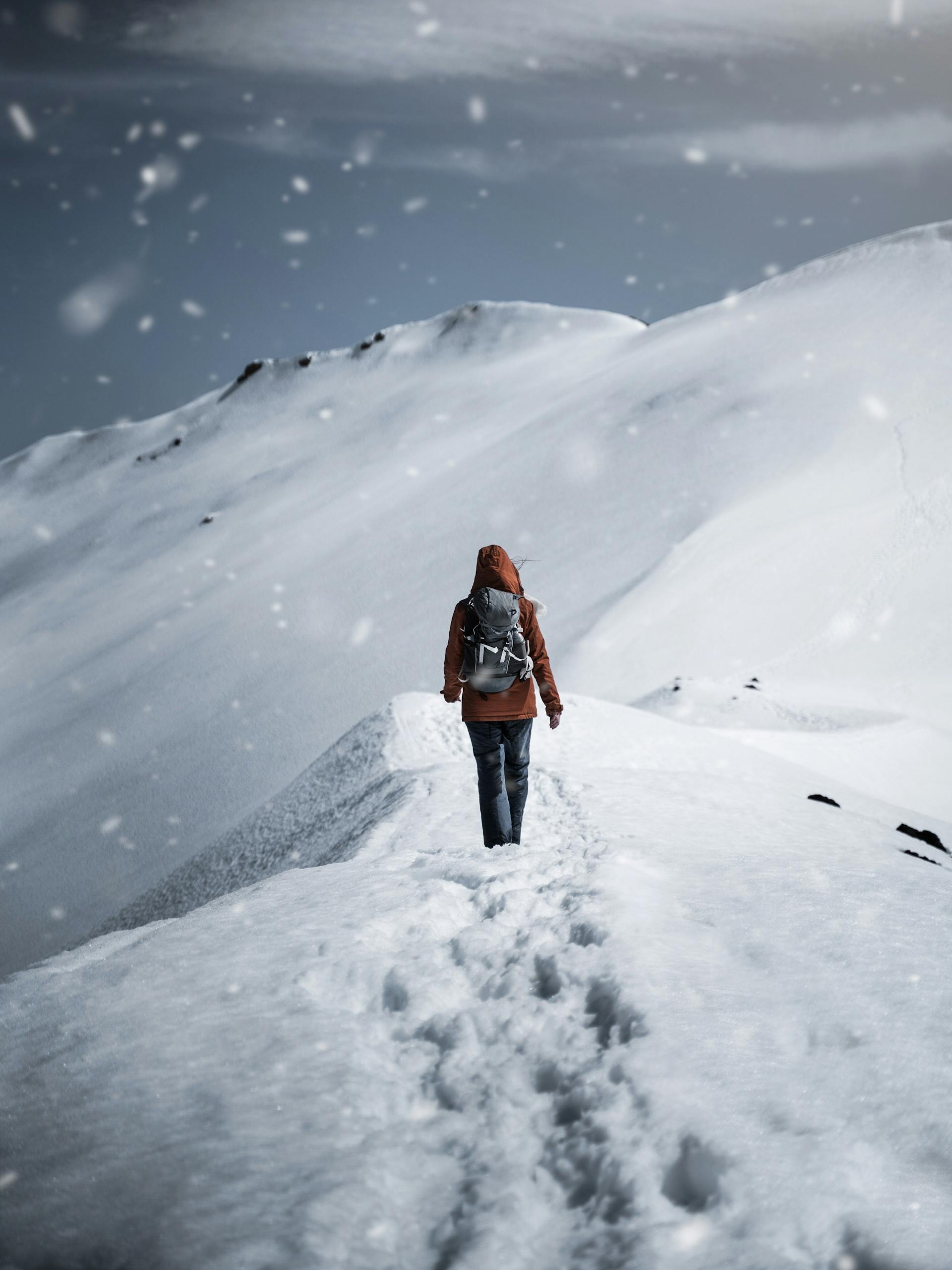 A lone hiker in a red jacket walks up a snow-covered mountain path during a snowfall. Photo by Gantas Vaičiulėnas