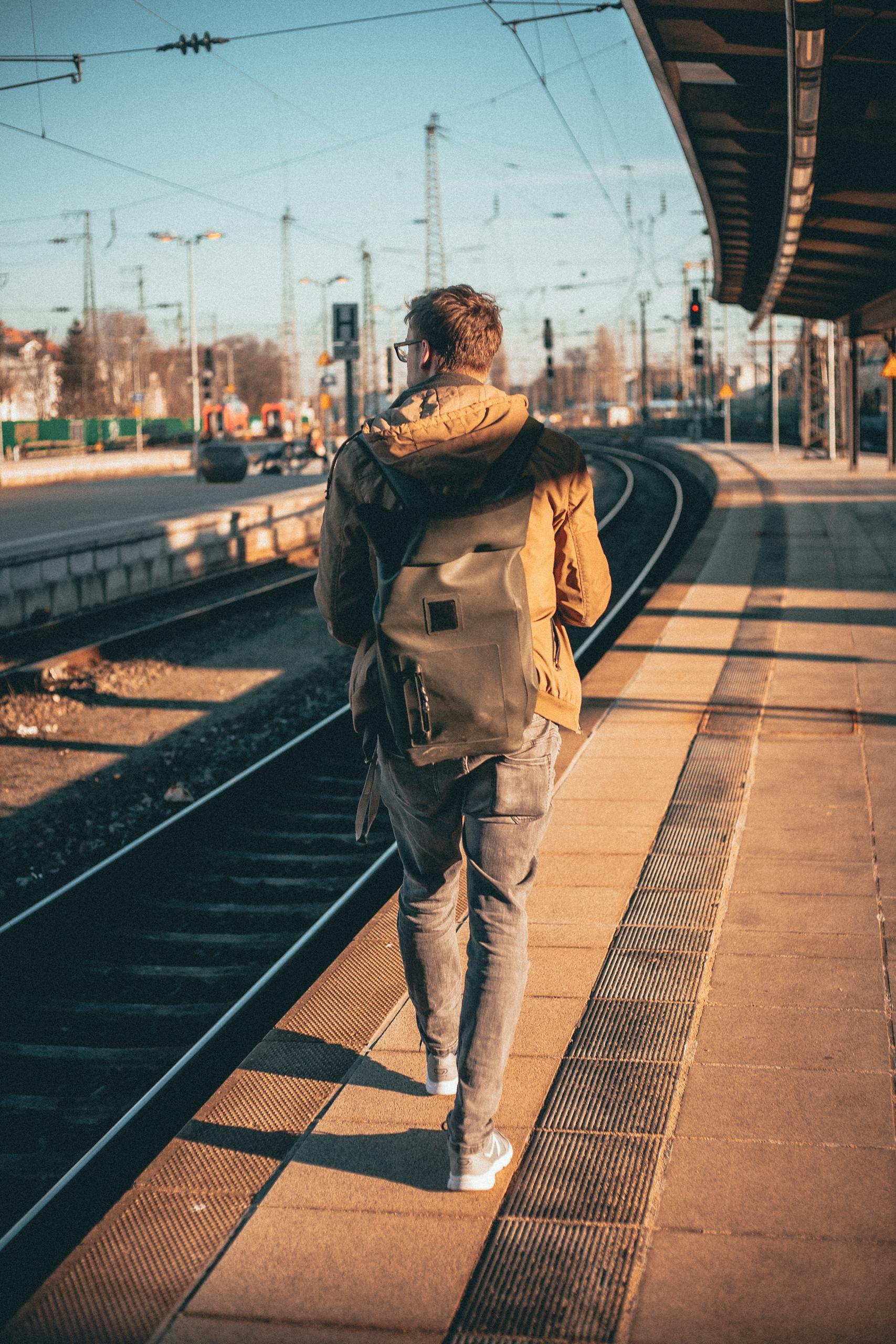 A man with a backpack walks along a train platform, capturing a journey moment in urban daylight. Photo by Malte Luk