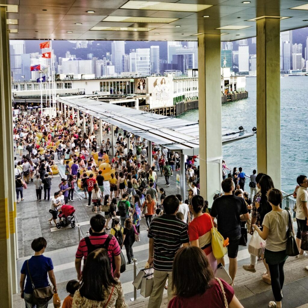 A vibrant crowd at the Hong Kong Island ferry terminal with a skyline backdrop.