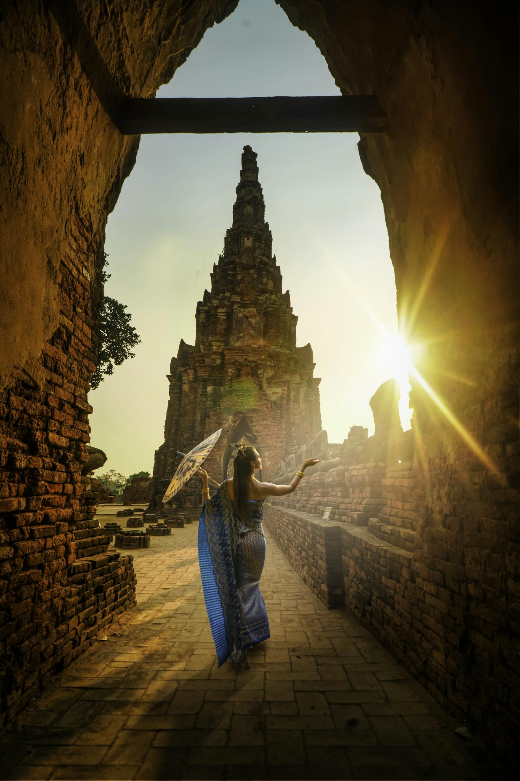 A woman in traditional wear stands with an umbrella at a historic Ayutthaya temple during sunset. Photo by Alex P