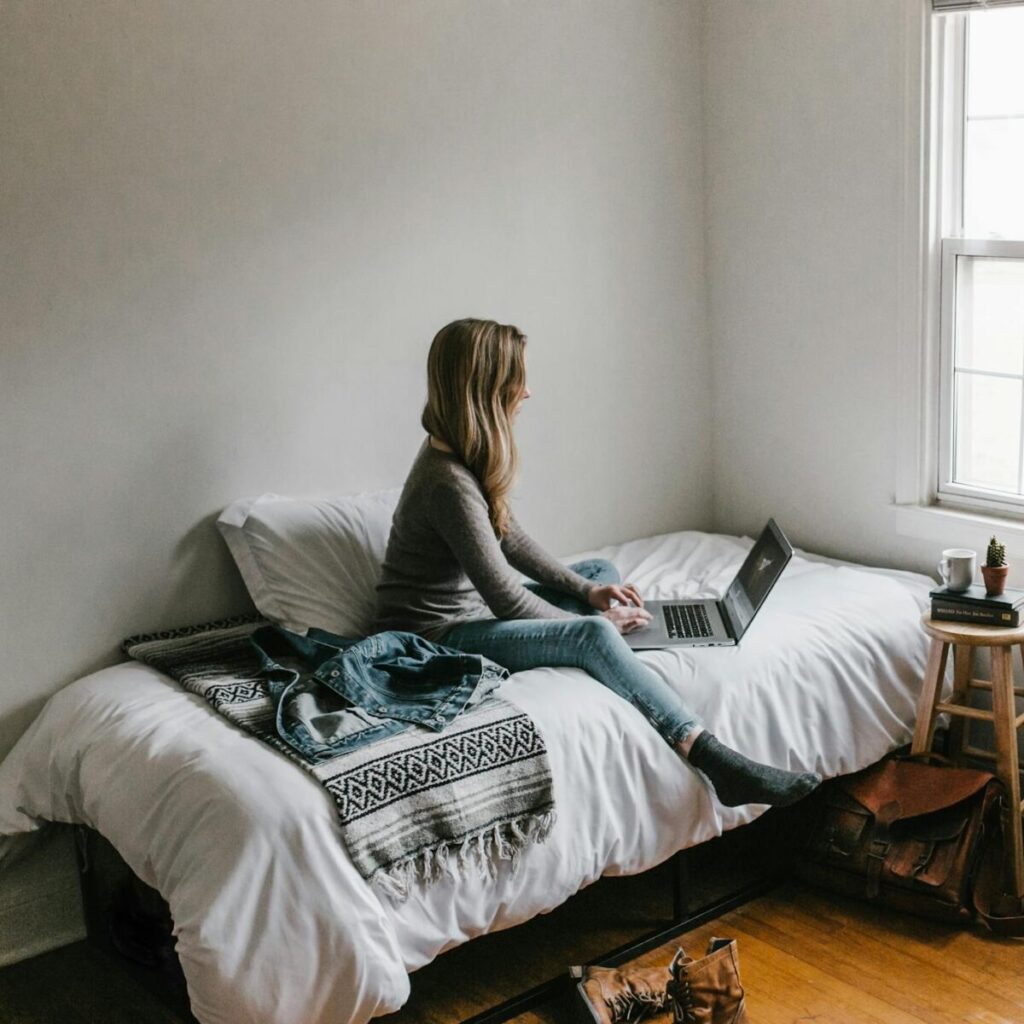 A young woman works on a laptop in a cozy, minimalist bedroom setting, showcasing remote work lifestyle.