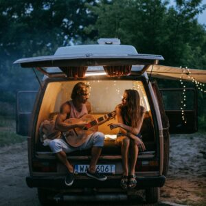 Photo by KoolShooters Two adults enjoying a musical road trip evening in a camper van, surrounded by nature.
