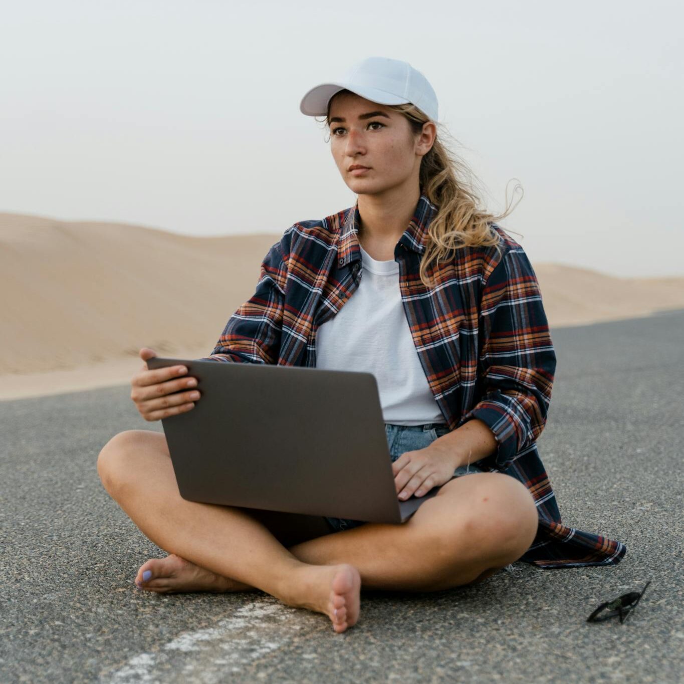 Young woman using laptop on a desert road, wearing casual attire and hat, symbolizing remote work.