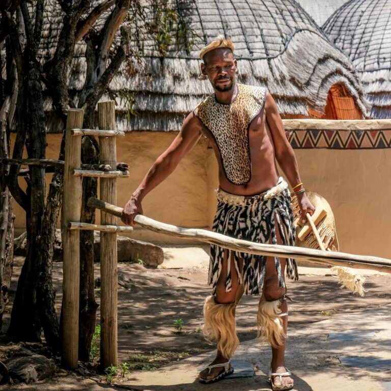 A man in traditional attire poses outdoors in a South African tribal setting.
