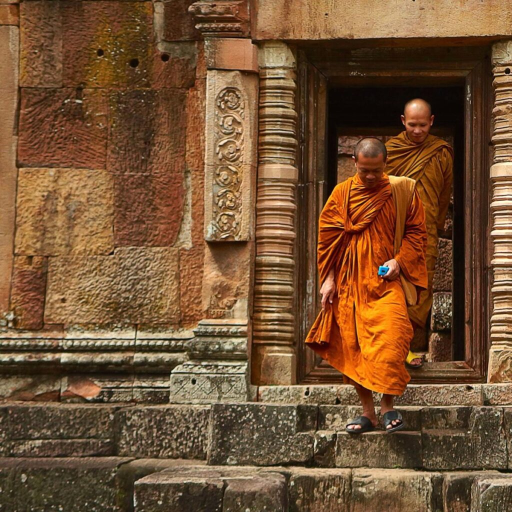 Buddhist monks in orange robes step out of an ancient stone temple in Thailand, showcasing historical architecture.