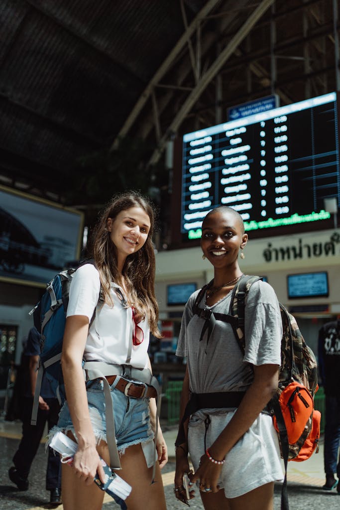 Two friends with backpacks smiling at a train station, ready to travel.