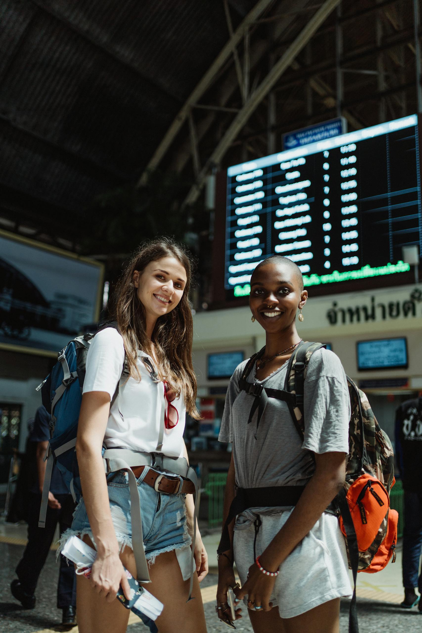 Two friends with backpacks smiling at a train station, ready to travel.