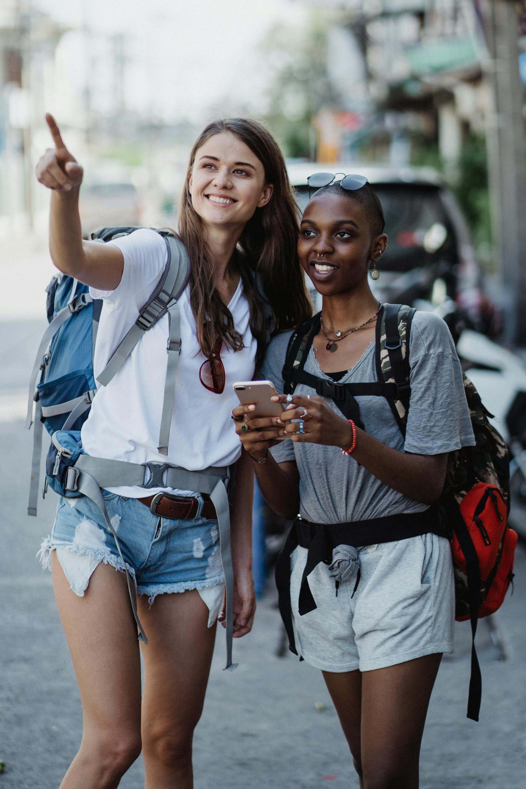 Two smiling women enjoying a day of sightseeing in an urban area, pointing and using a smartphone to navigate.