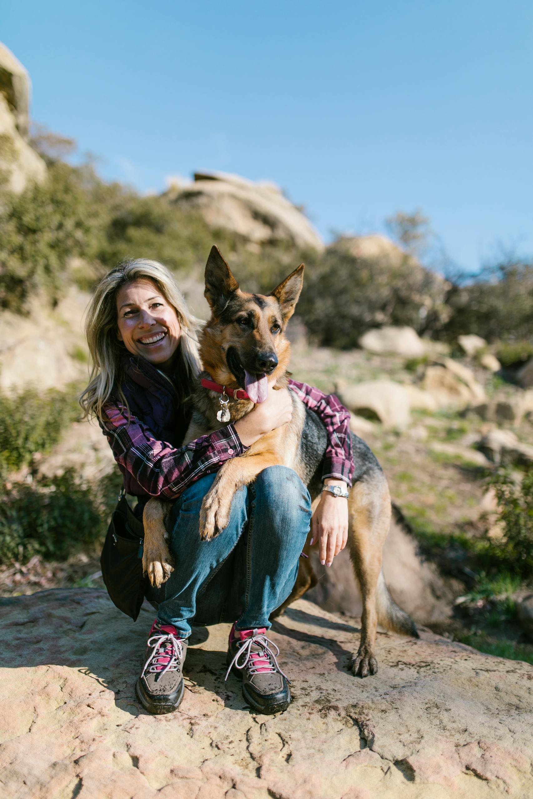 A smiling woman embraces her German Shepherd during a sunny day hike.