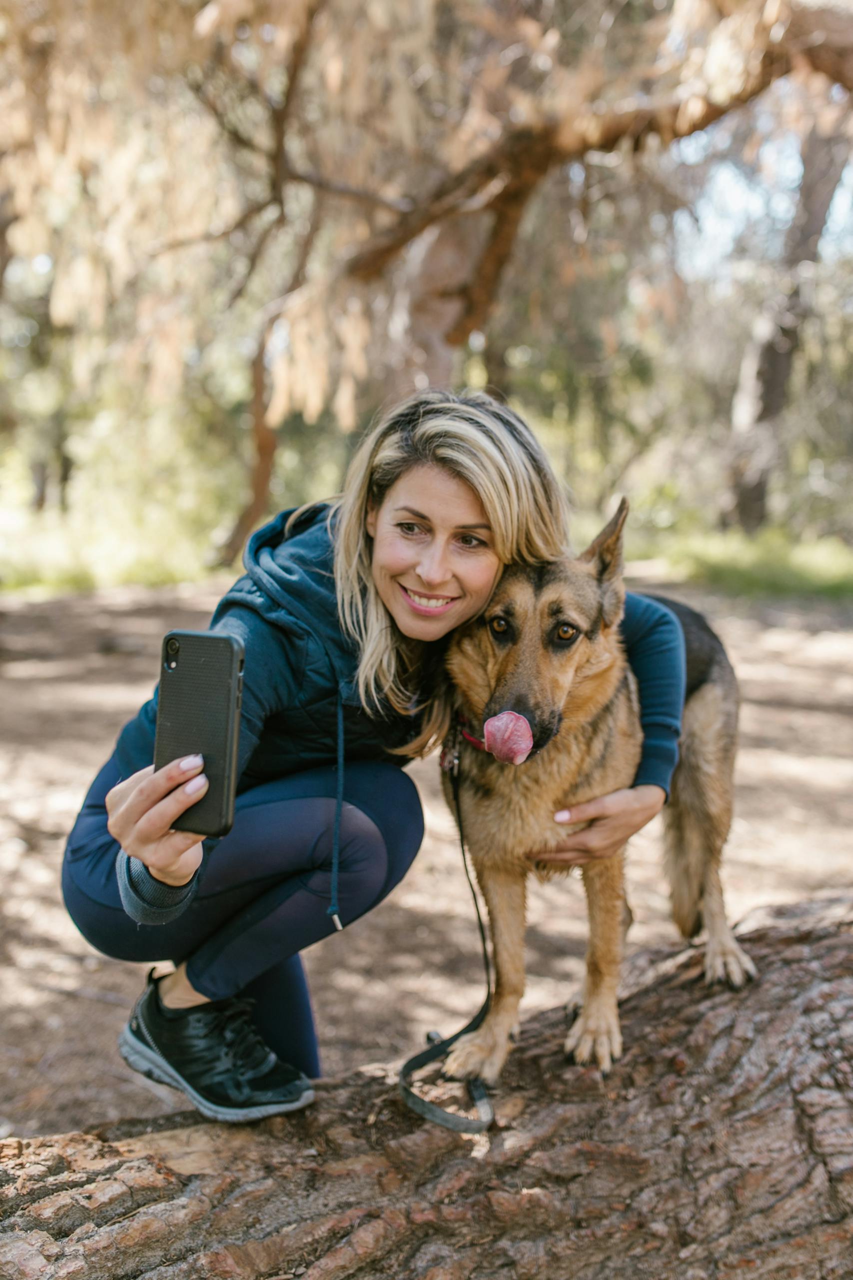 A woman in a blue jacket takes a selfie with her German Shepherd in a forest setting.