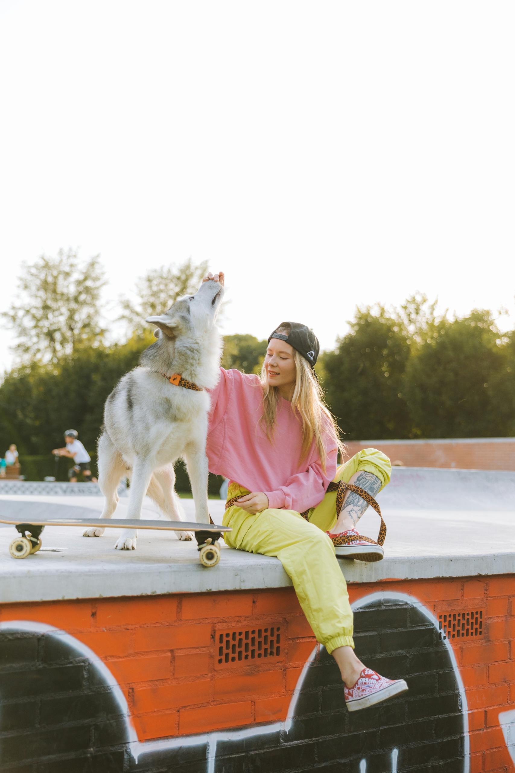 A woman in a pink shirt and cap sits at a skate park with her husky on a sunny day.