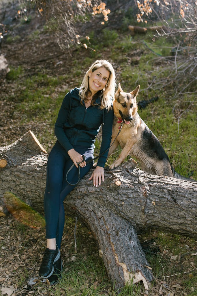 A woman sits with her German Shepherd on a tree trunk in a sunny forest clearing.