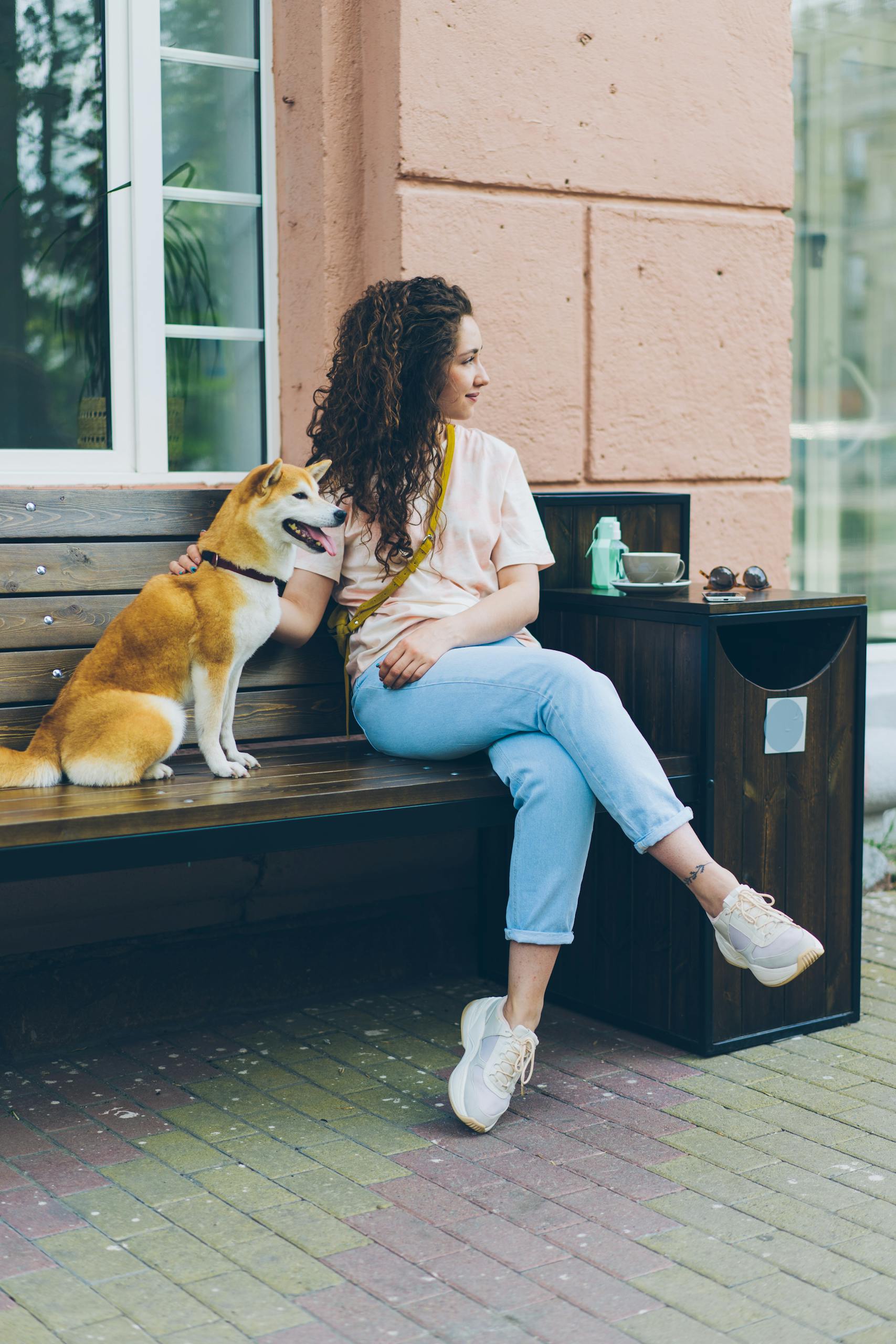 Woman with curly hair sits on a bench with a Shiba Inu dog, enjoying a relaxed urban atmosphere.