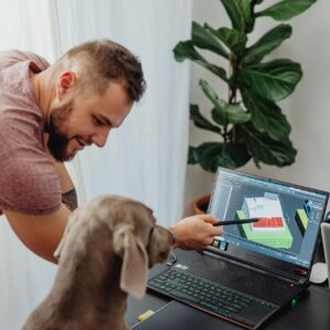 A man instructs a dog using a laptop in a home office with a potted plant nearby.