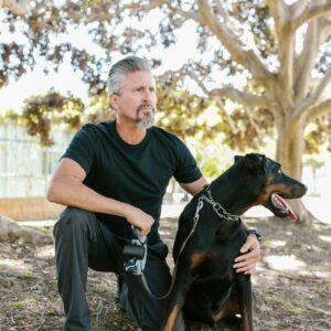 A man kneeling beside a Doberman dog in a sunlit park, showcasing companionship and calmness.