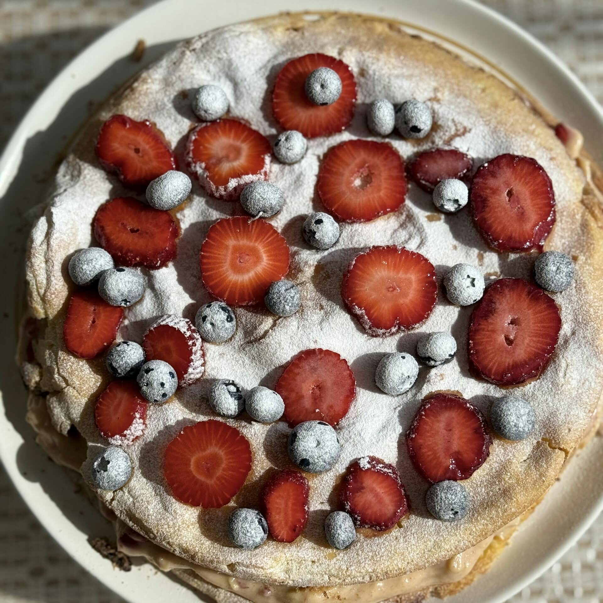 Delicious meringue cake topped with fresh strawberries and blueberries, dusted with powdered sugar.