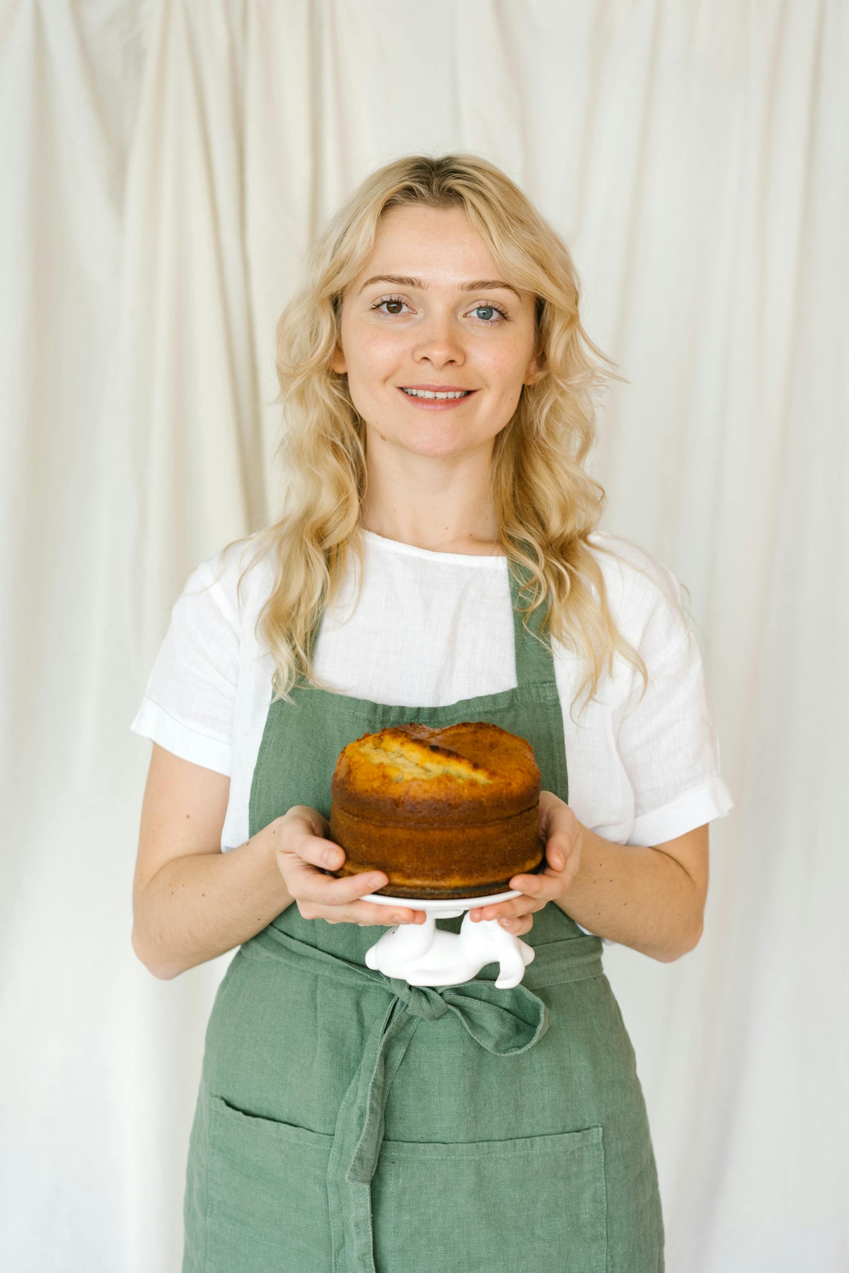 Smiling woman in green apron holds a freshly baked cake against a neutral white background.