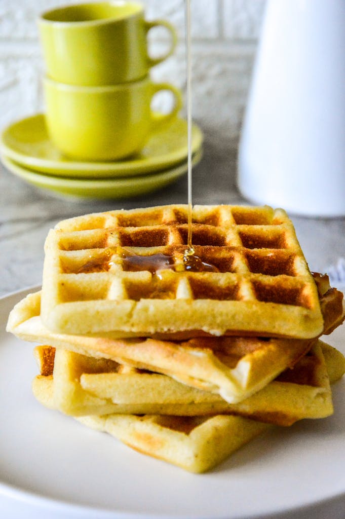 Stack of golden waffles with syrup pouring indoors with green cups.