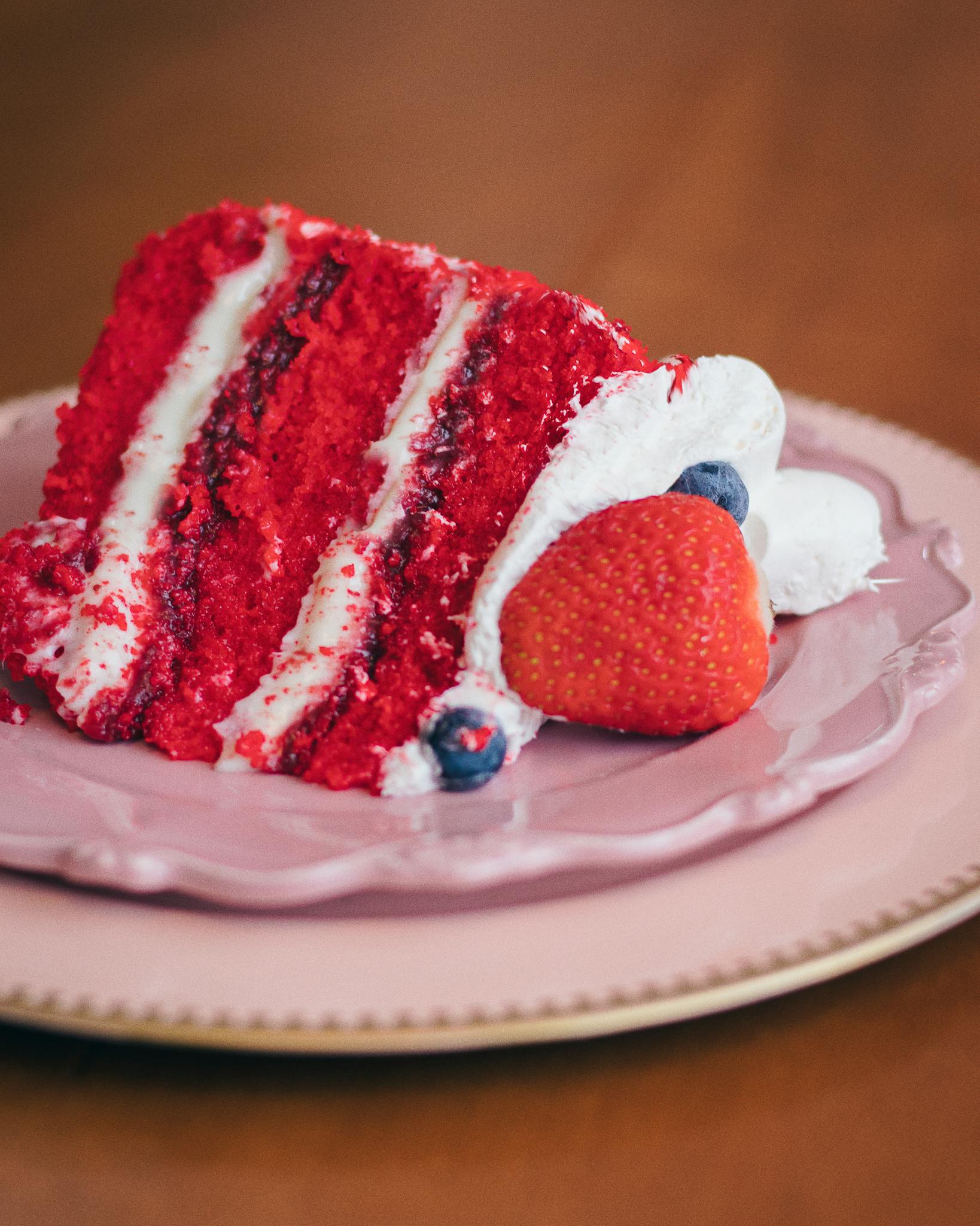 Sumptuous red velvet cake slice with whipped cream and berries on a pink plate.