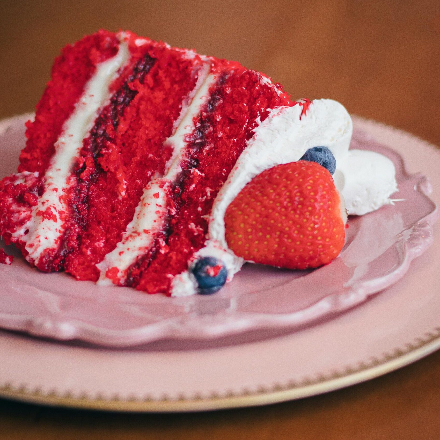 Sumptuous red velvet cake slice with whipped cream and berries on a pink plate.