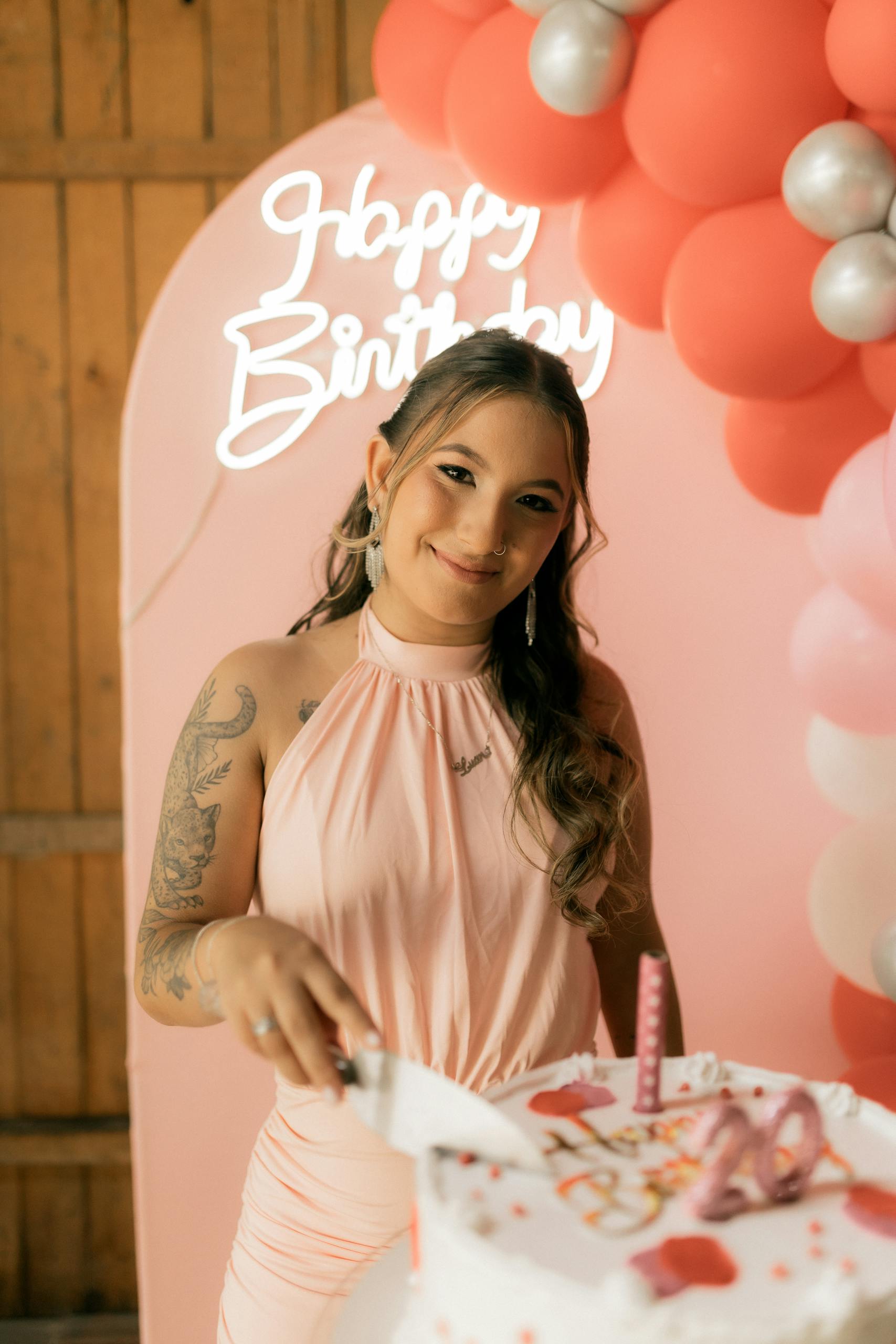 A woman cutting a birthday cake in a festive setting with balloons and a 'Happy Birthday' sign.