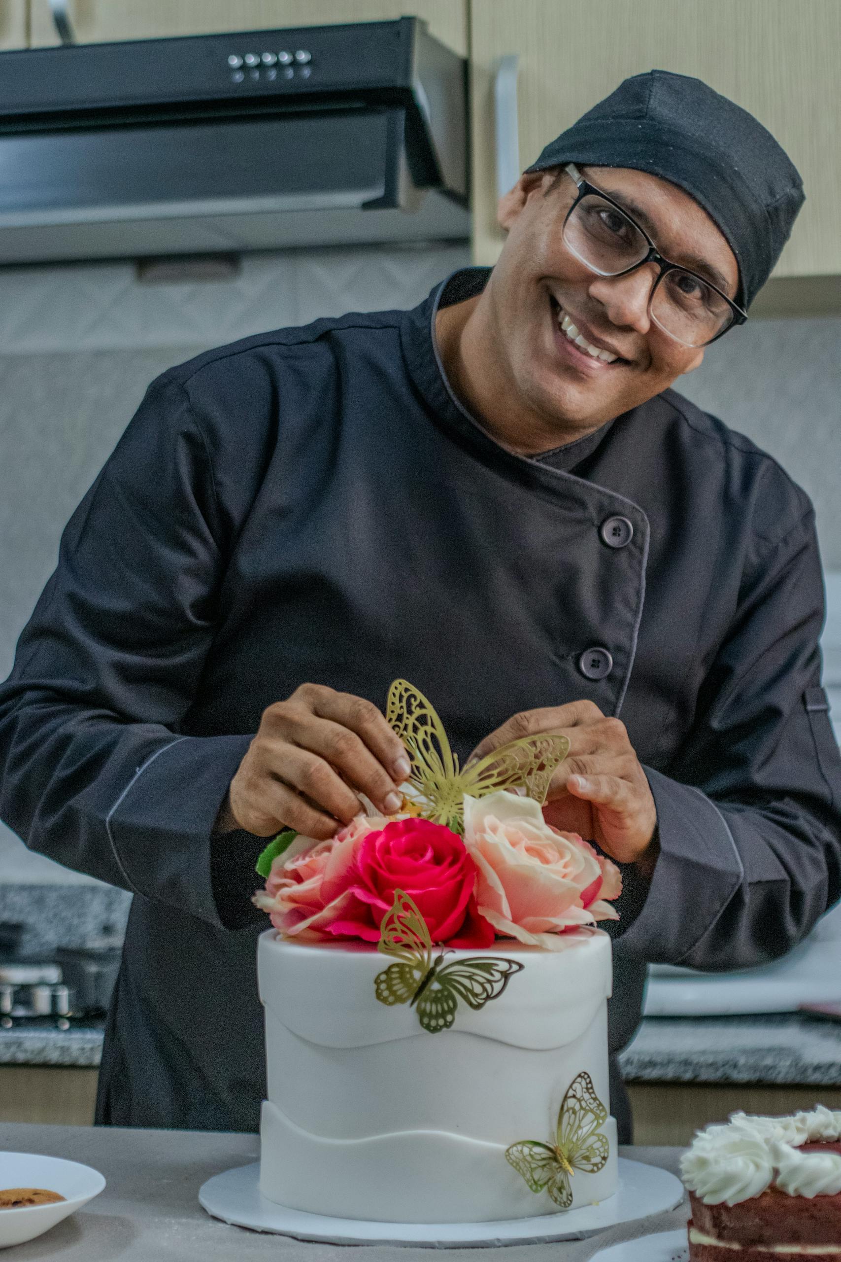 Chef in a black uniform decorates a white cake with roses and butterflies in a modern kitchen.