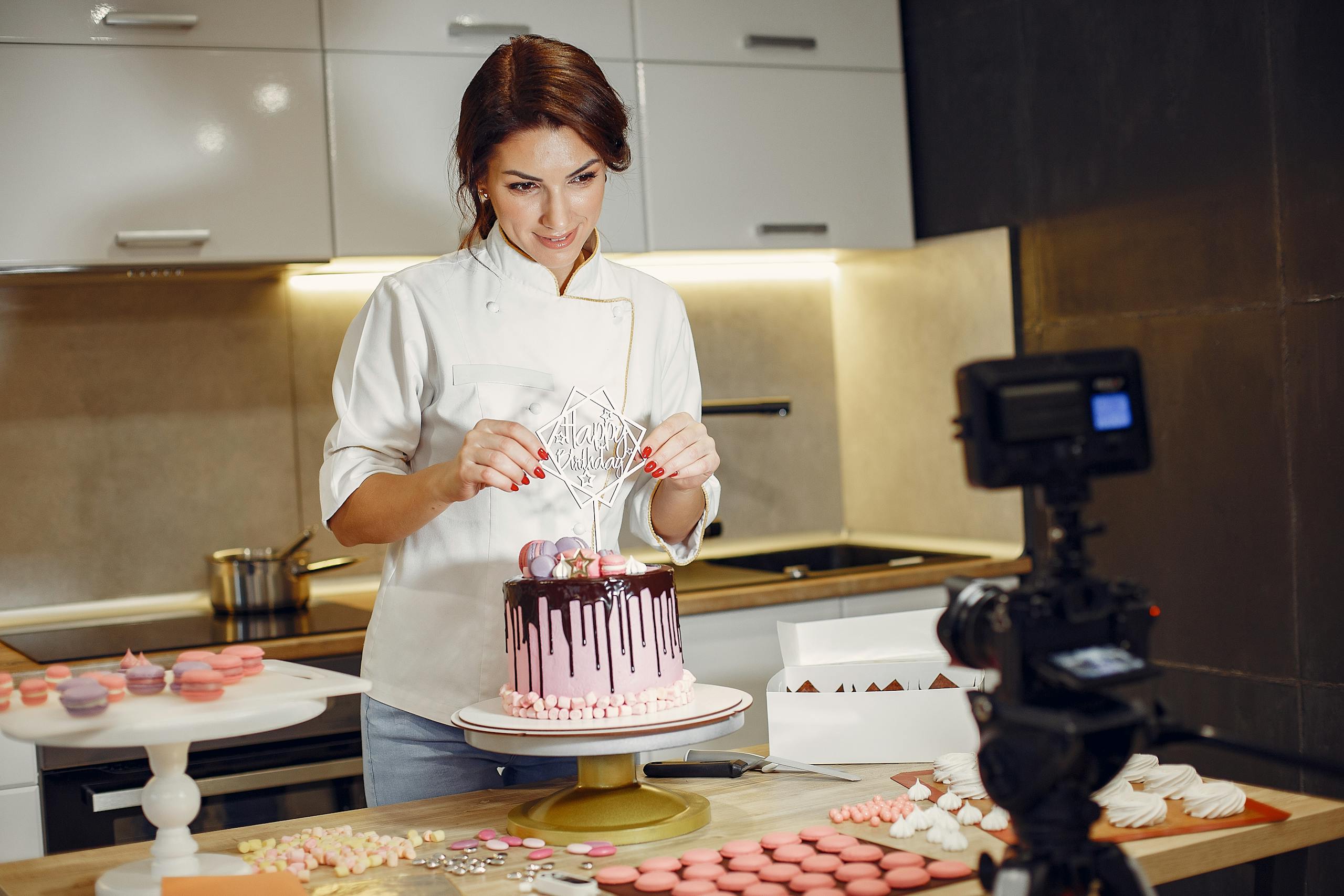 Smiling female pastry chef in uniform decorating cake and looking at lens while standing in front of video camera in modern bakery