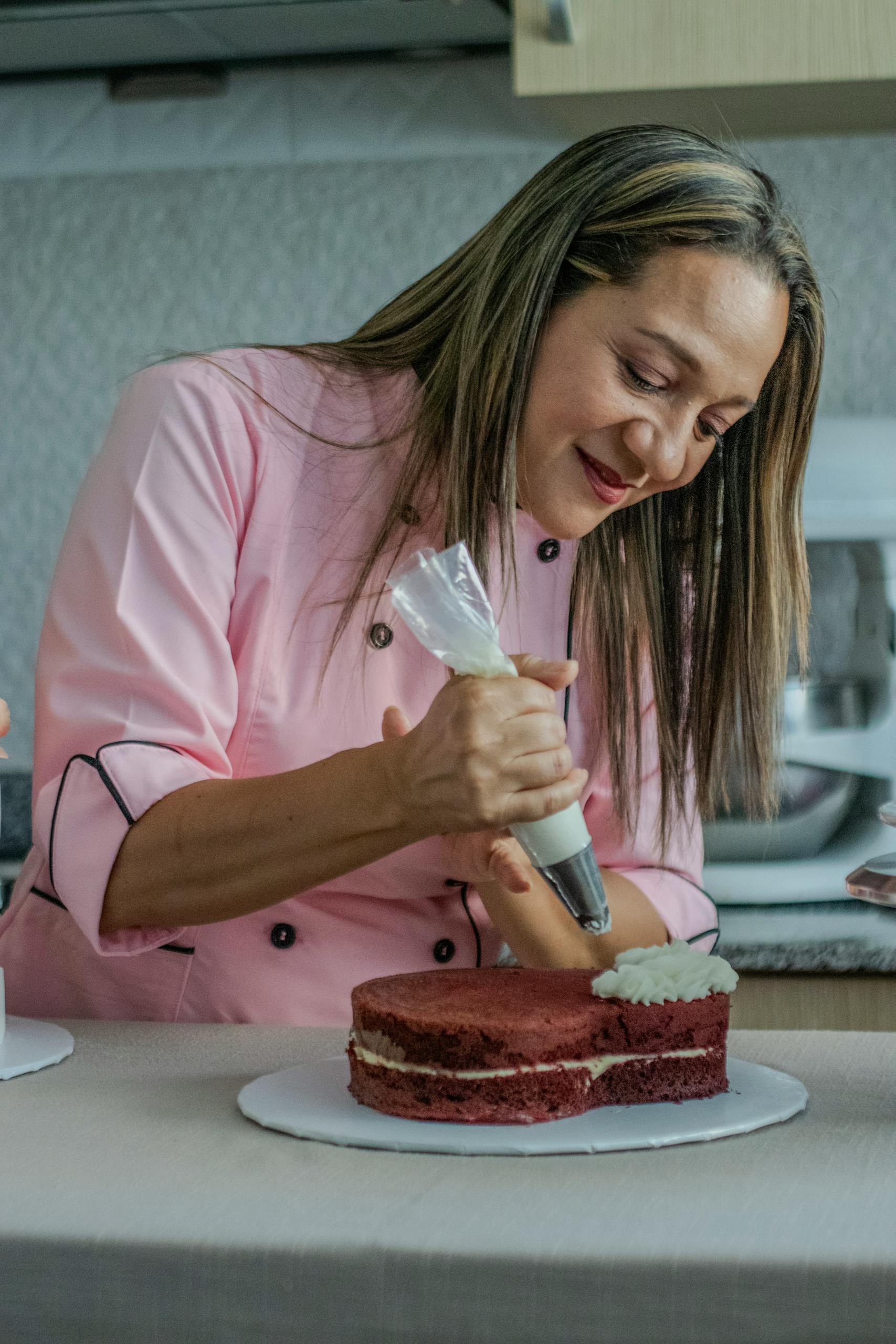 Woman in a pink chef jacket skillfully decorates a cake with frosting in a kitchen setting.